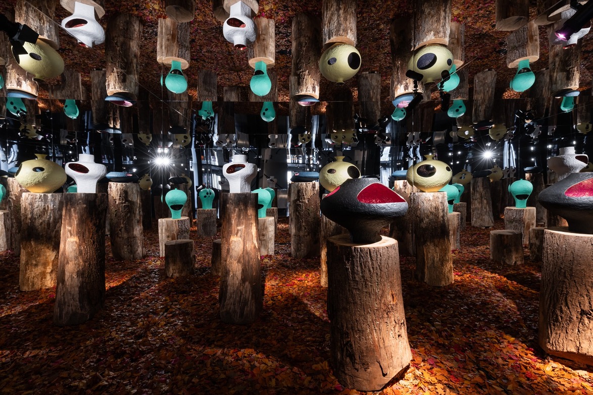 Art installation featuring colorful abstract sculptures on tree stumps, surrounded by mirrored walls and autumn leaves.
