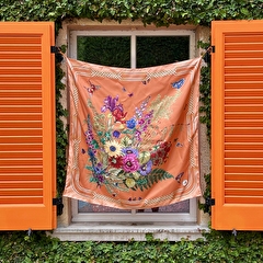 Orange floral scarf with various flowers hanging on a window with orange shutters, surrounded by green ivy.