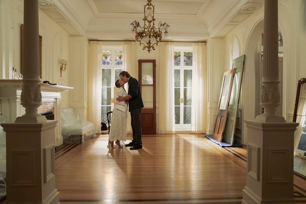 A couple embraces in a warmly lit, elegant room with wooden floors and stained glass windows.
