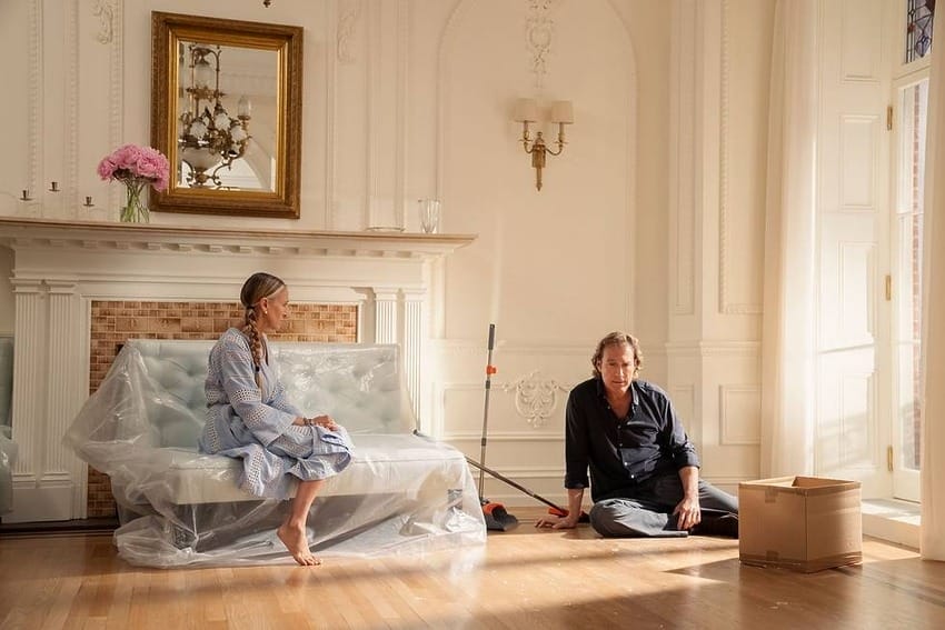 Two people sitting on the floor in a partially furnished room with a couch and unpacked boxes.