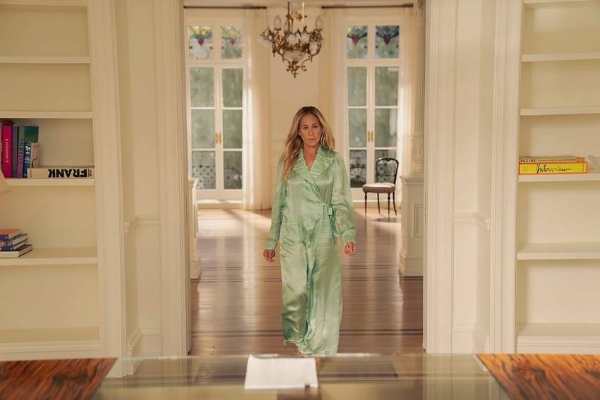 Woman in a light green robe walking through a stylish, well-lit room with bookshelves and a chandelier.