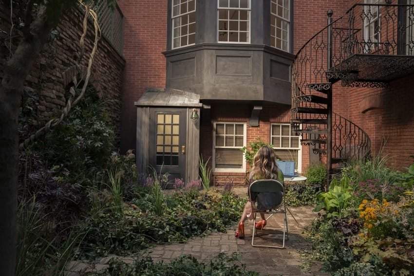 Woman sitting on a chair in a lush garden courtyard, working on a laptop, surrounded by brick walls and spiral staircase.