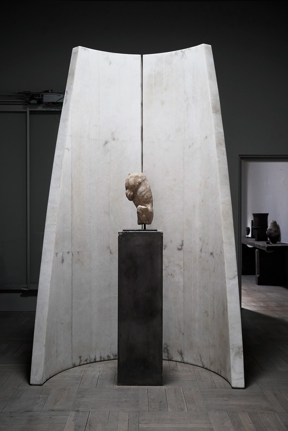 Marble sculpture on a dark pedestal framed by tall curved white marble panels in a dimly lit modern gallery space