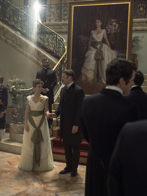 Elegant ballroom scene with people in vintage attire, a large portrait on the wall, and grand staircase in the background.