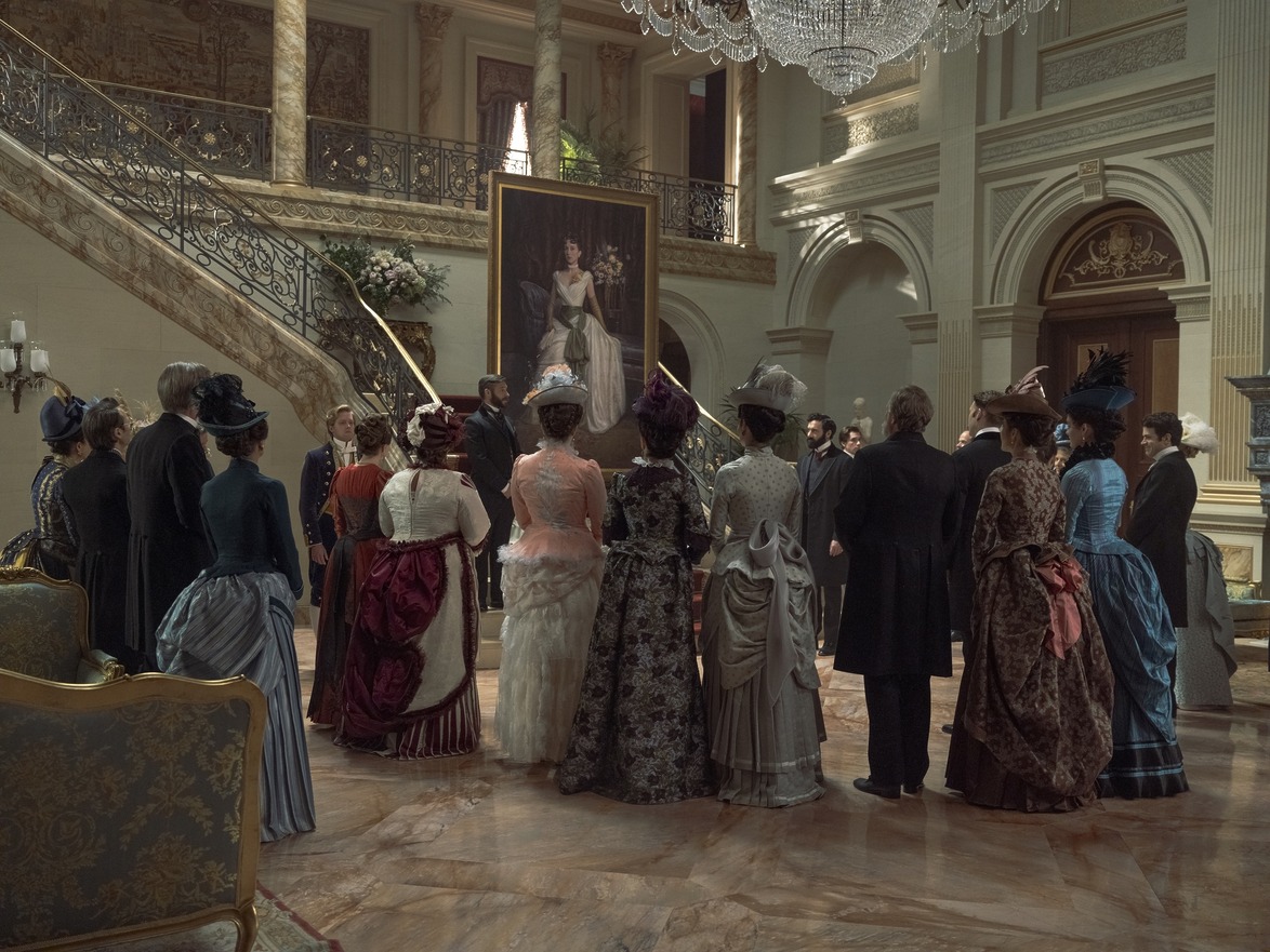Victorian-era gathering in an elegant mansion with guests in period clothing and a large portrait on the wall.