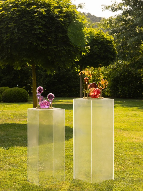 Two tall clear pedestals with decorative purple and red glass sculptures set outdoors in a lush green garden.
