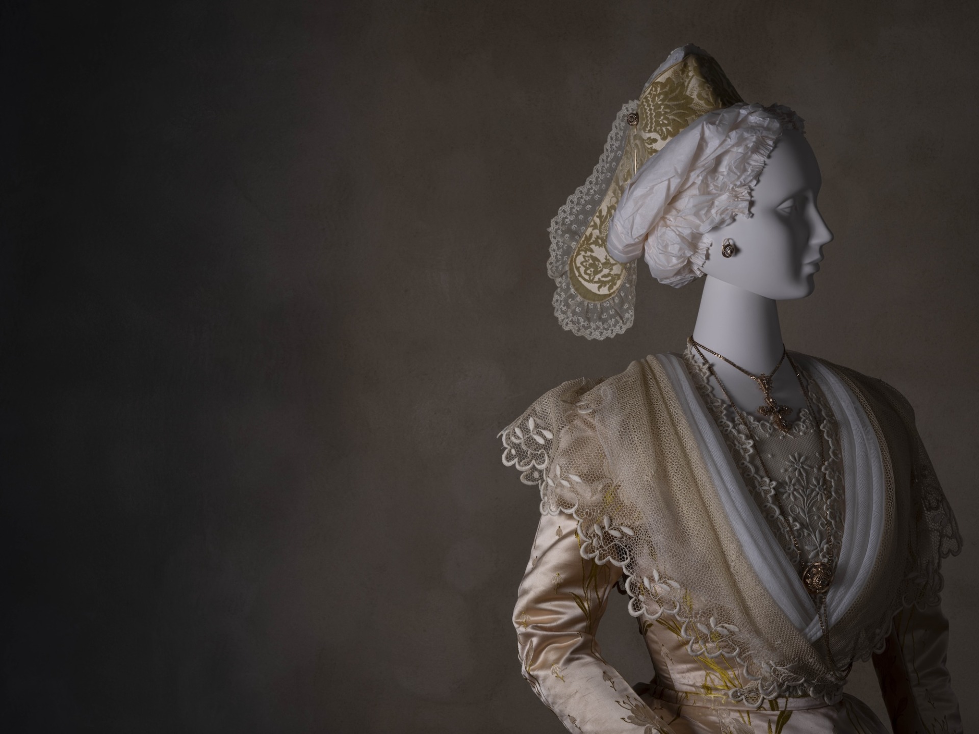 Mannequin in vintage attire with ornate lace headdress and jewelry against dark background.