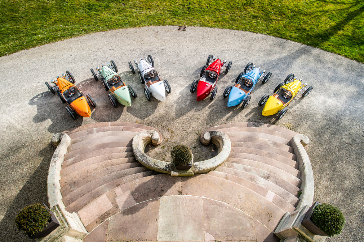 Six colorful vintage-style three-wheeled cars parked in a semicircle on a gravel path near ornate steps and greenery.