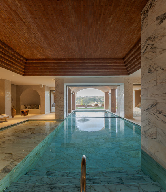 Indoor swimming pool with marble tiles, wooden ceiling, and scenic view through open doors leading to a balcony with hills.