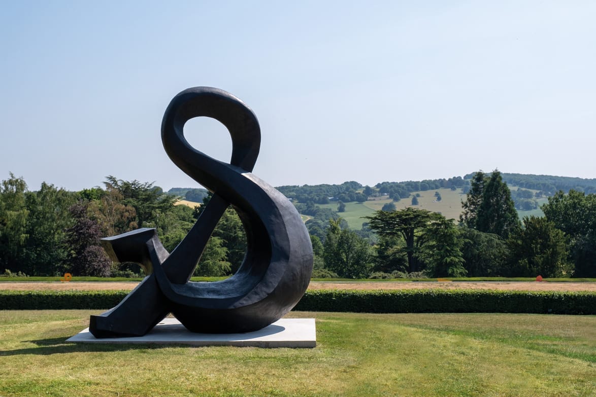 Large black ampersand sculpture in a grassy field with rolling hills and trees in the background.