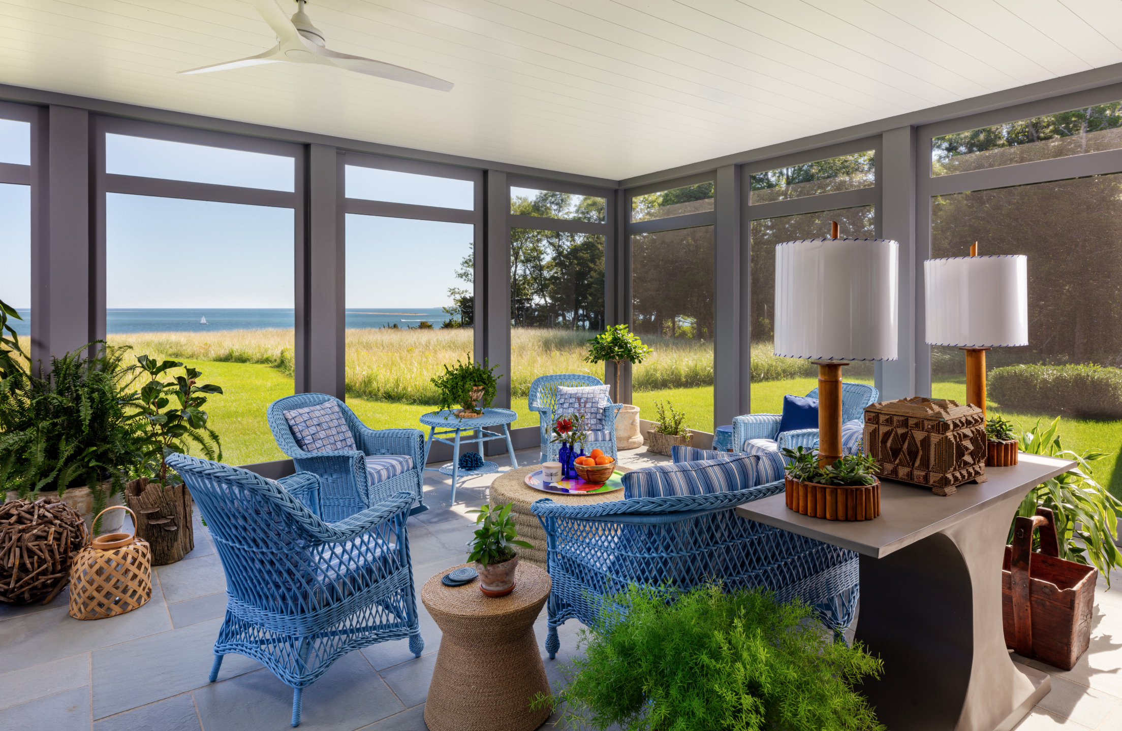 Cozy sunroom with blue wicker furniture, sea view, and lush greenery, capturing a serene summer atmosphere.