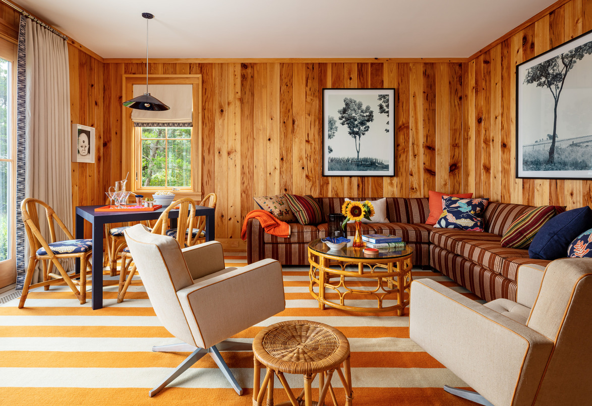 Cozy living room with wood-paneled walls, striped rug, colorful sofa, table, chairs, and framed artwork.