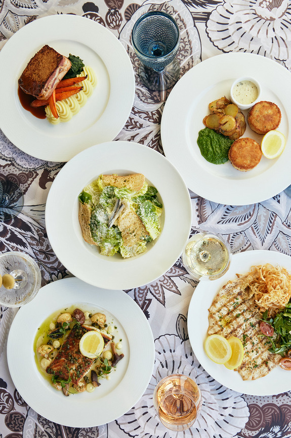 Five elegant plated dishes on a table with varied savory foods and garnishes, accompanied by glasses of wine and water.