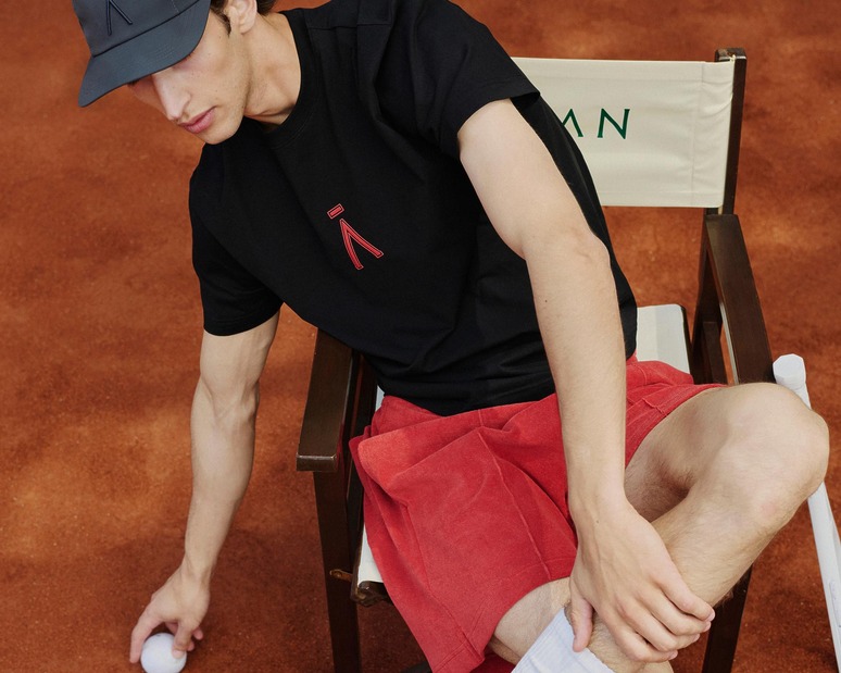 Man in black t-shirt and red shorts sitting on a chair, holding a tennis ball on a clay tennis court.