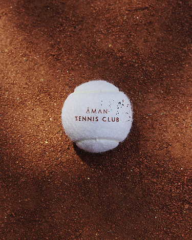 Tennis ball with "Aman Tennis Club" branding on a clay court surface.