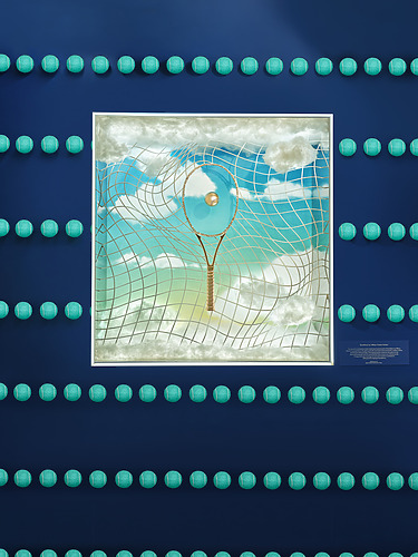 Framed artwork of a clouded sky with a tennis racket pattern, surrounded by tennis balls on a blue wall.
