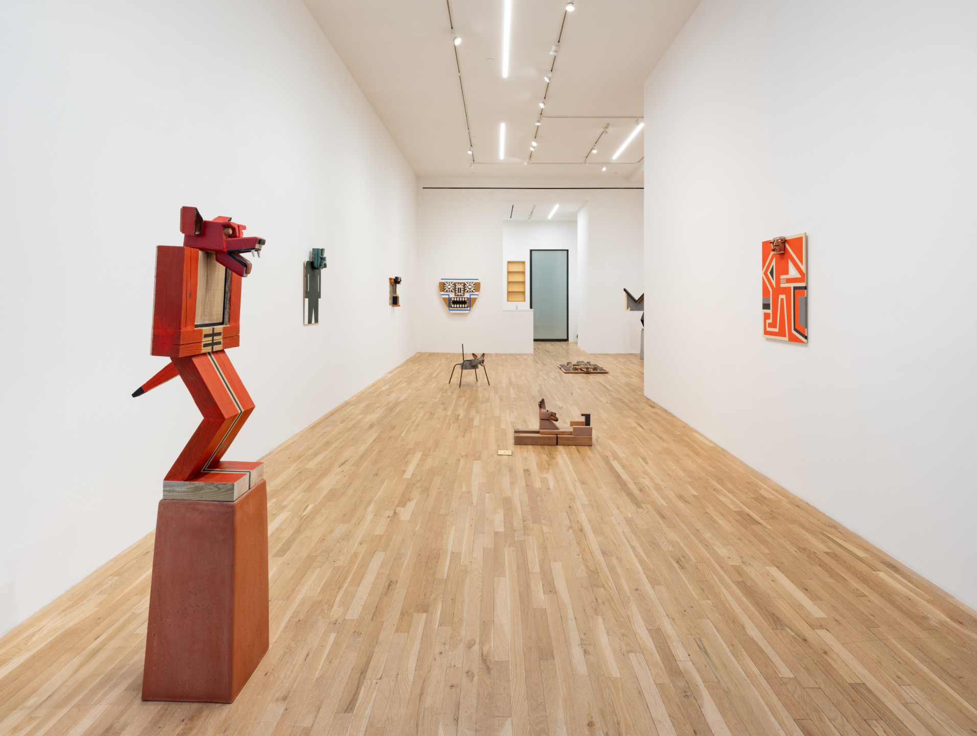 Art gallery with wooden sculptures and abstract paintings displayed on a wooden floor and white walls.