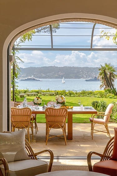 View of outdoor dining area with table and chairs, overlooking a lawn, sea, sailboat, yachts, and distant landscape through an archway.