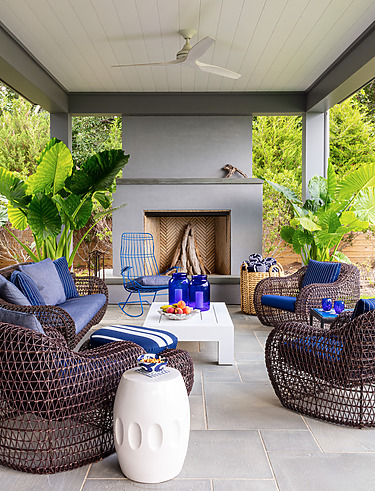 Outdoor patio with dark wicker furniture, blue cushions, a white table, a fireplace, and lush green plants surrounding the space.