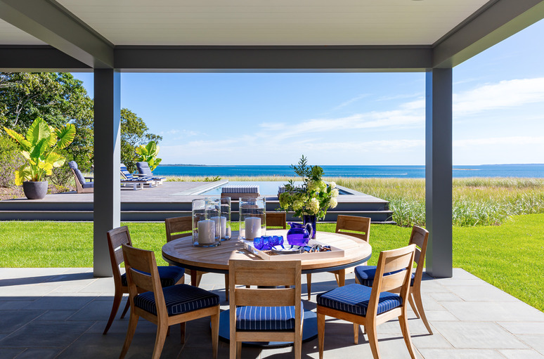 Outdoor patio with round dining table and ocean view in the background, surrounded by greenery and deck chairs.