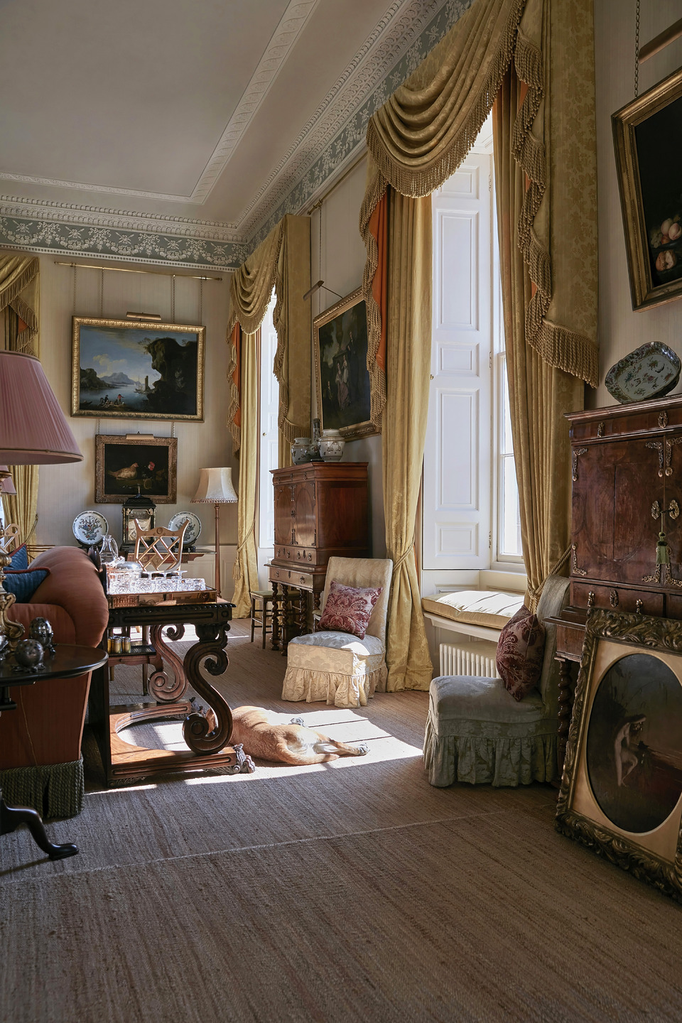 Elegant vintage living room with antique furniture, large windows, ornate drapes, and a dog lounging in sunlight.
