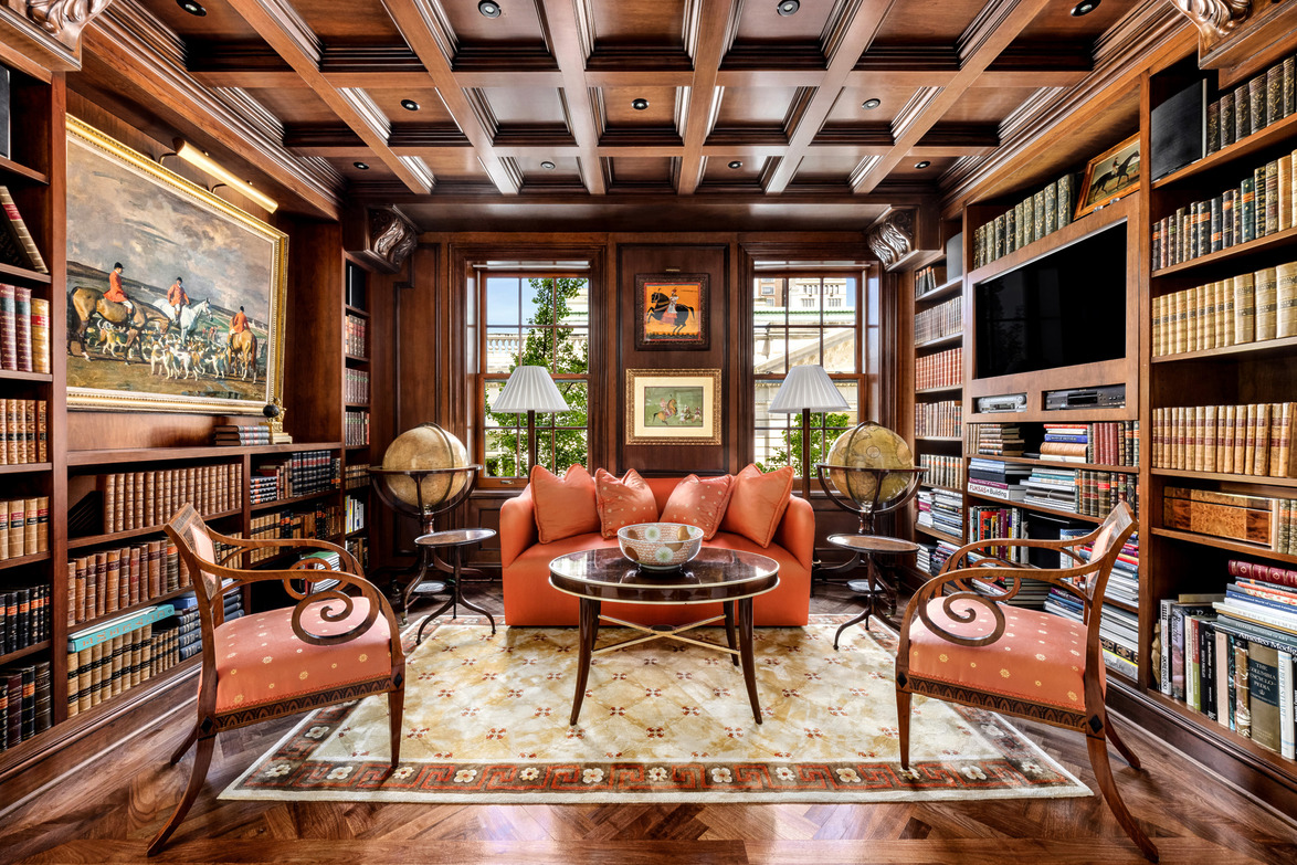 Elegant home library with bookshelves, ornate ceiling, orange sofa, two globe tables, art, and large windows.
