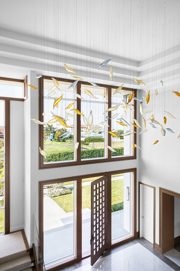 Colorful glass leaves hang from a high ceiling in a modern open foyer with large windows and a wooden door.