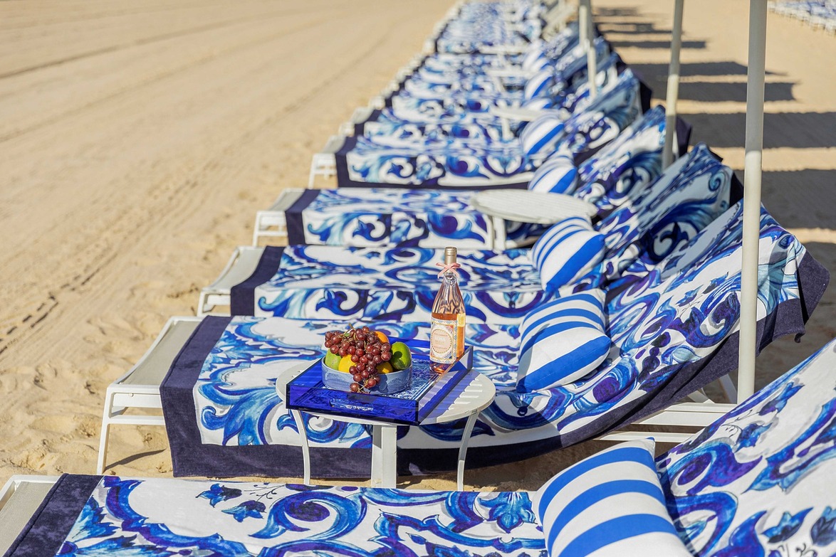 Beach lounge chairs with blue patterned towels, a tray with fruit, and a bottle of rosé wine on a sandy beach.