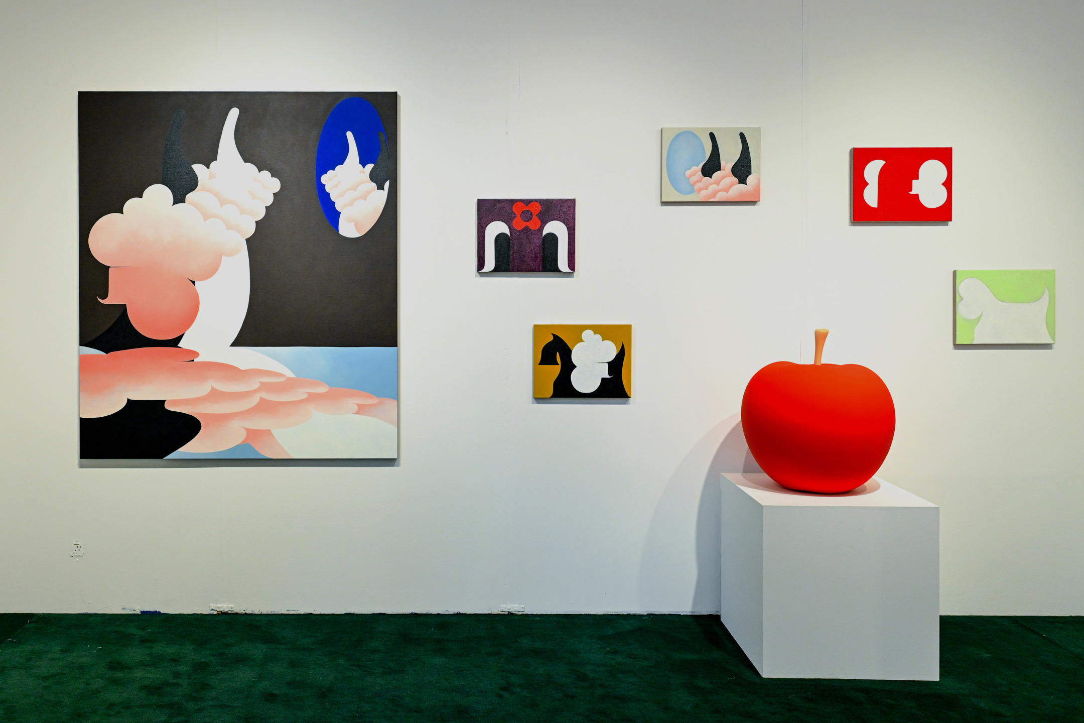 Art gallery display with colorful abstract paintings and a large red apple sculpture on a pedestal against a white wall.