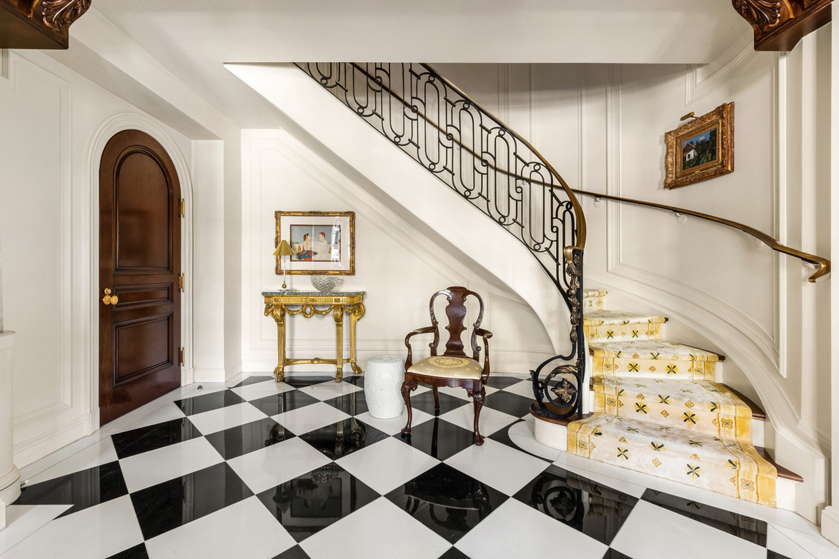 Elegant foyer with grand staircase, checkered floor, ornate table, and classic chair, all lit by natural sunlight.