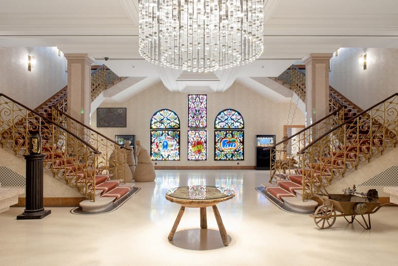 Elegant hotel lobby with stained glass windows, chandelier, dual staircases, and a round table in the center.