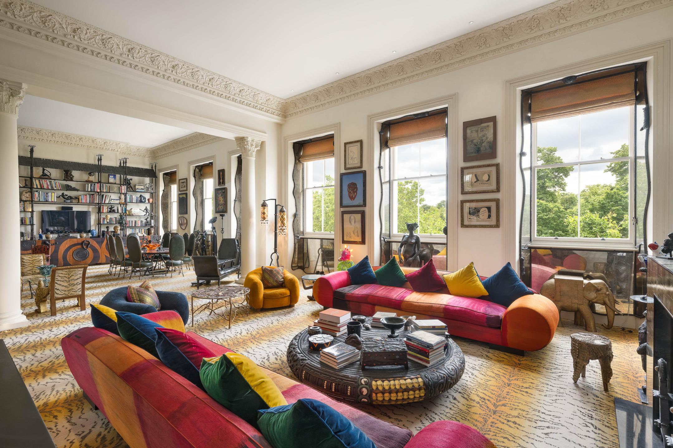 Colorful living room with rainbow sofas, eclectic decor, large windows, and views of green trees outside.