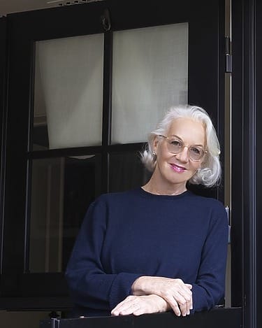 Elderly woman with white hair and glasses smiling while standing by a modern black door with curtains in the background.