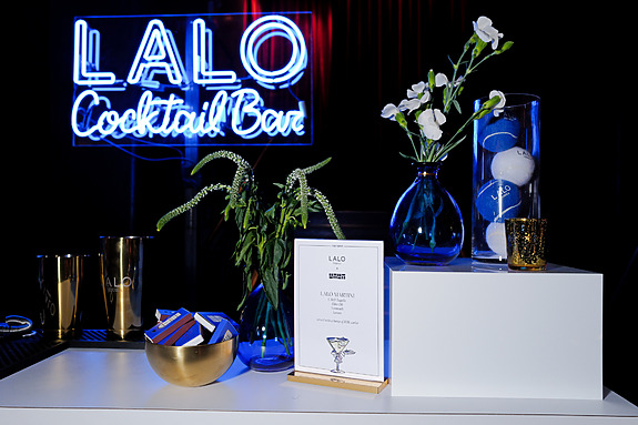 Neon sign reading "LALO Cocktail Bar" above a bar setup with flowers, menu, gold shakers, and tennis balls in a vase.