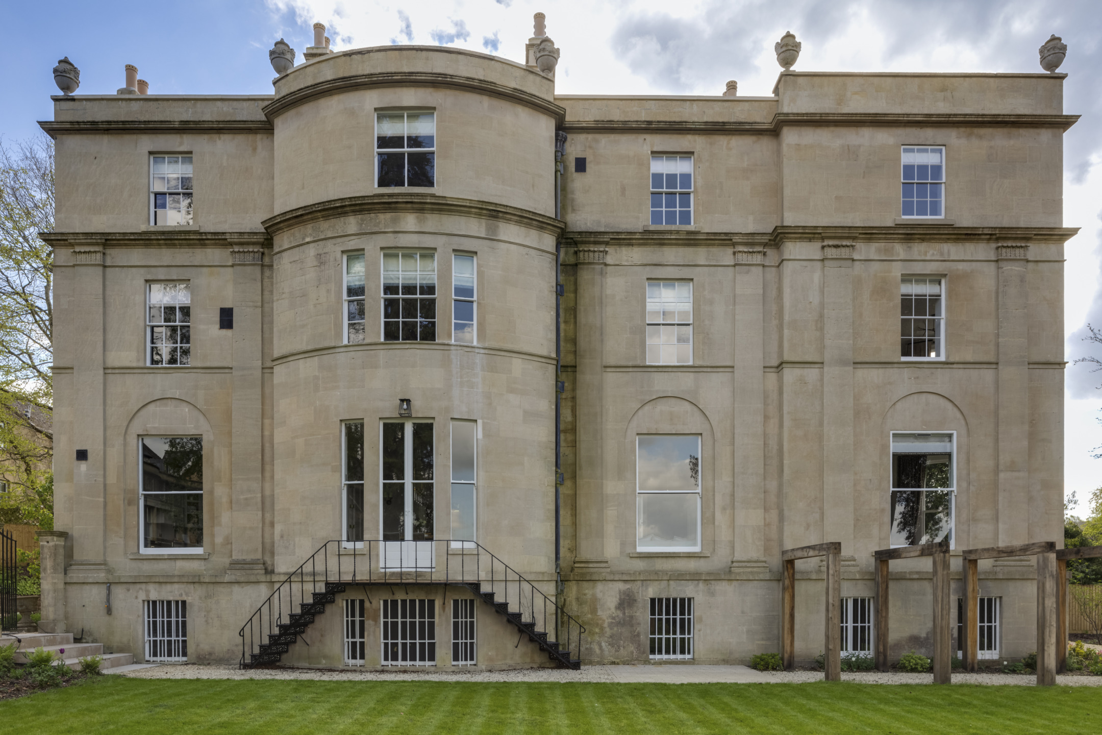 Historic stone building with symmetrical windows, central staircase, and manicured lawn under a partly cloudy sky.
