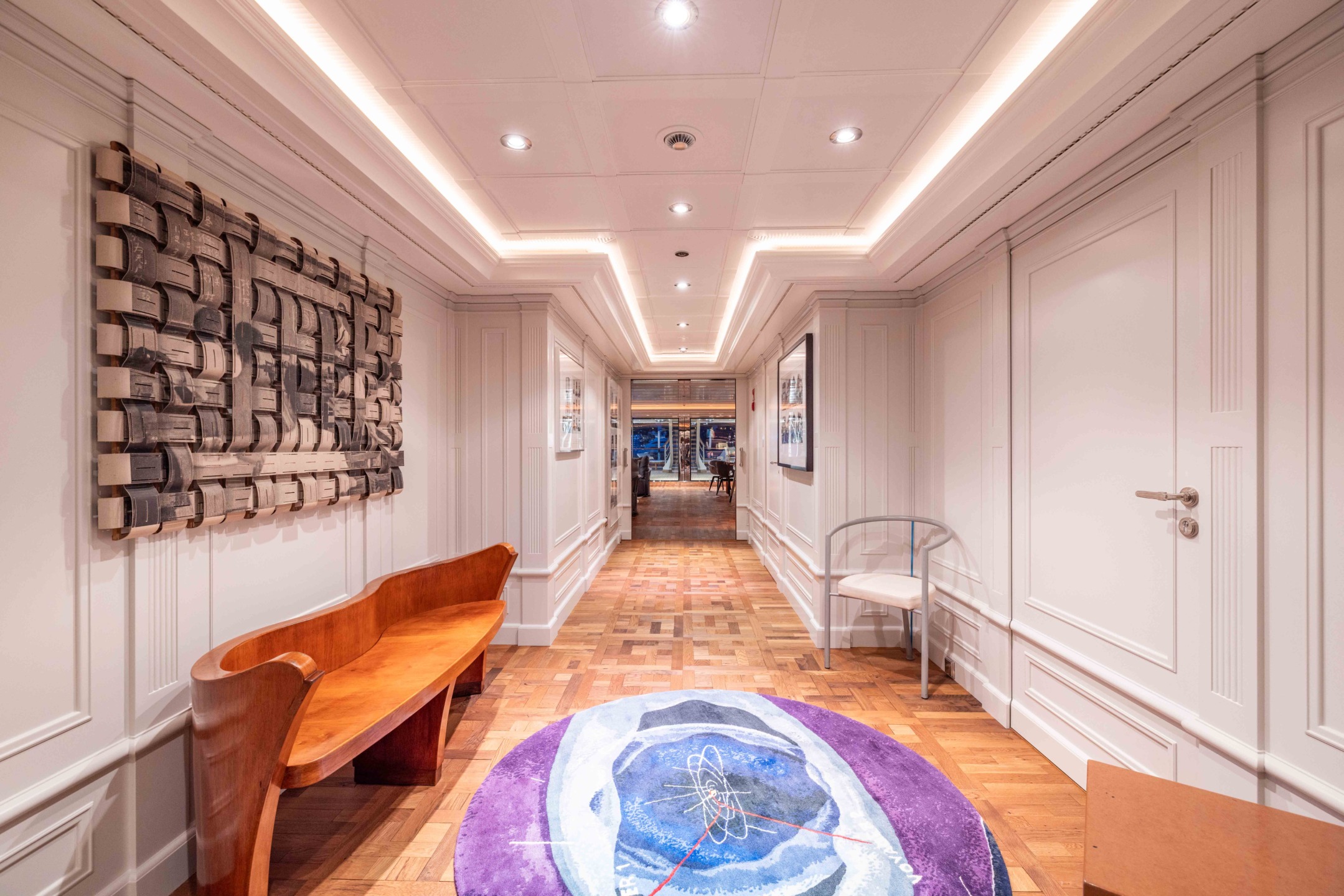 Elegant hallway with parquet flooring, modern art on walls, wooden bench, and a purple rug leading to a bright room.