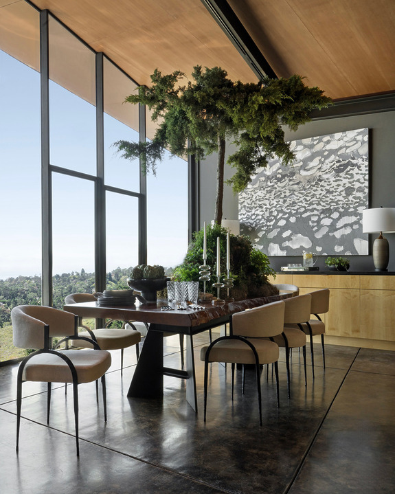 Modern dining room with large windows, sleek furniture, and a central table centerpiece featuring a small tree arrangement.