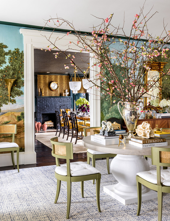 Elegant living room with green chairs, large floral centerpiece, and a view into a dining area with a chandelier.