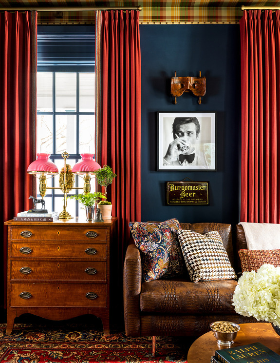 Cozy living room with dark walls, red curtains, vintage furniture, patterned pillows, and framed black-and-white photo on wall.