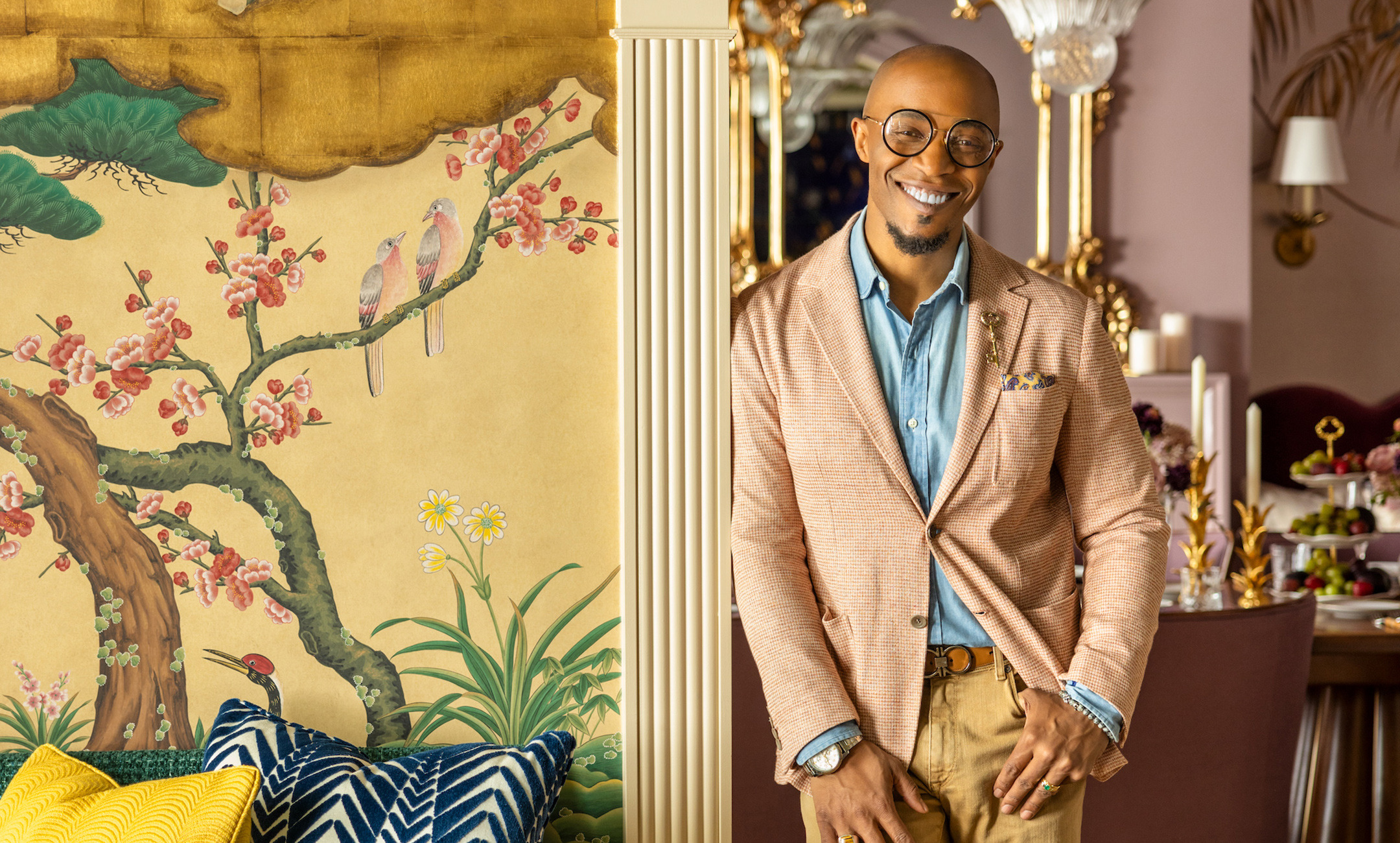 Person smiling in a stylish room with vibrant wallpaper, colorful cushions, and elegantly set dining table in the background.