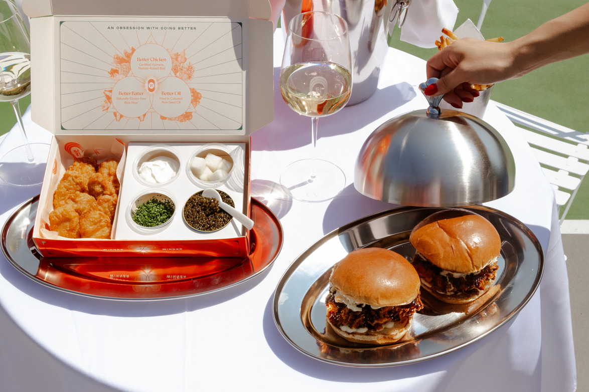 Open box with fried chicken bites, dips, two burgers on a platter, a hand lifting a cloche, and glasses of white wine on a table.