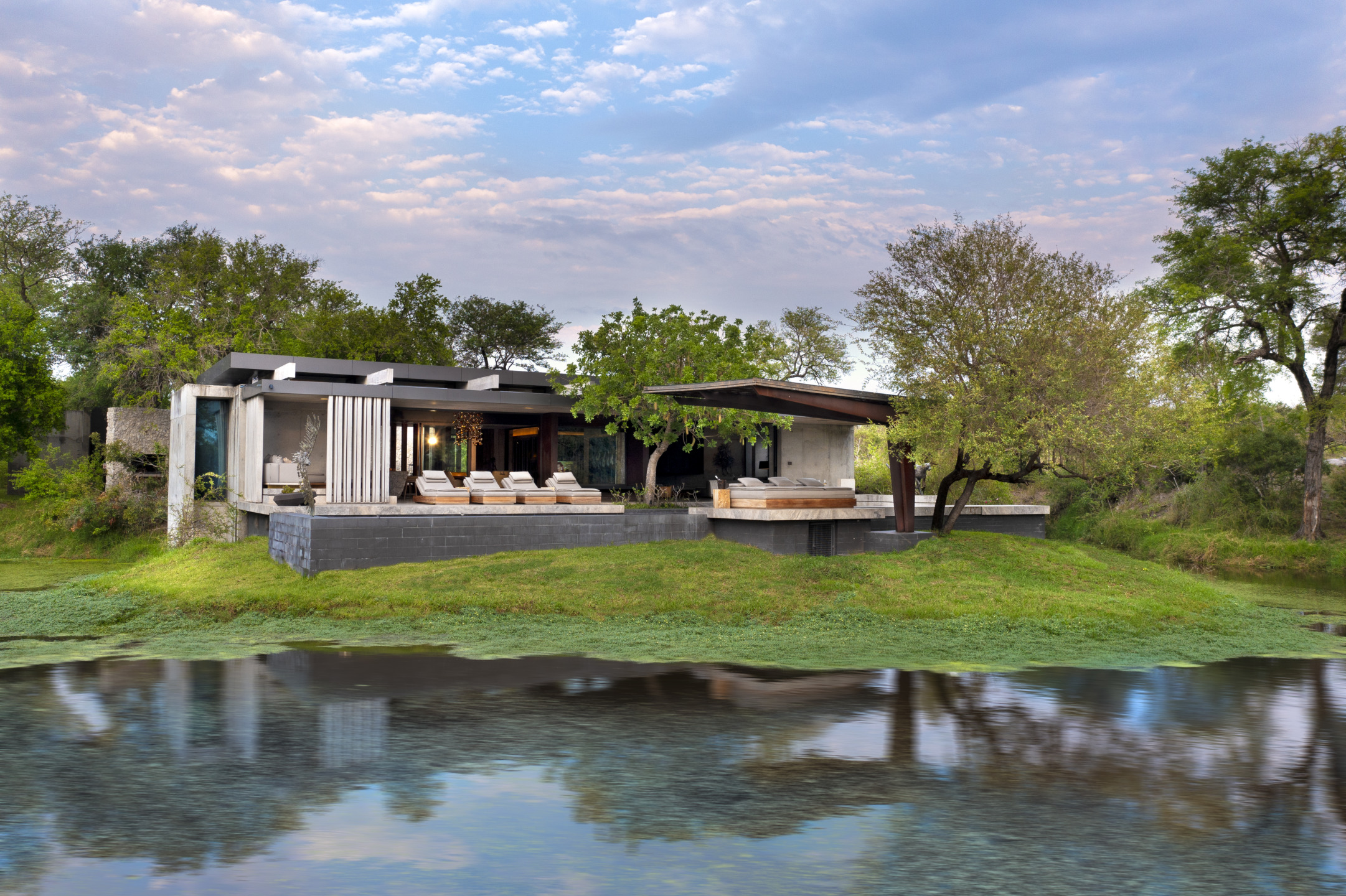 Modern lakeside house with large windows, surrounded by trees and greenery, reflecting on calm water under a cloudy sky.