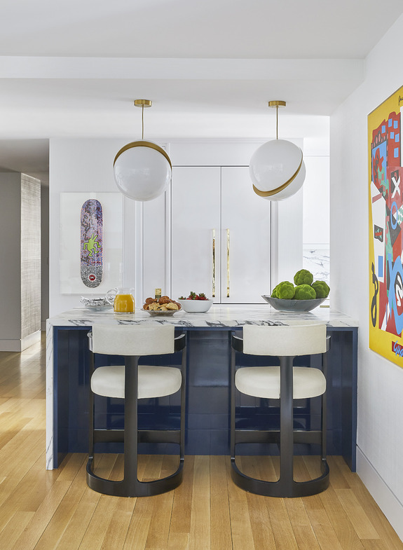Modern kitchen with a marble island, two chairs, hanging lights, and colorful wall art.