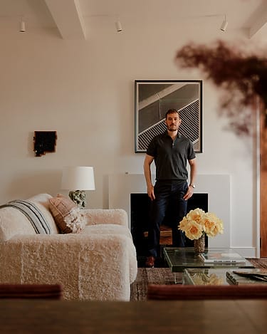Man standing in a modern living room with a sofa, glass table, flowers, and artwork on the wall.