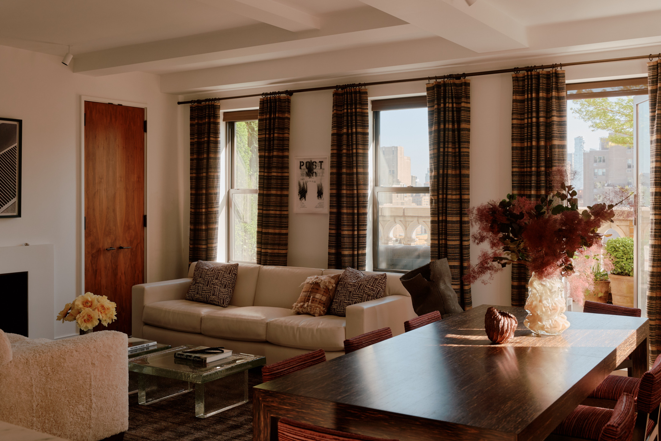 Sunlit living room with a beige sofa, plaid curtains, a wooden table, and glass coffee table in a modern apartment setting.