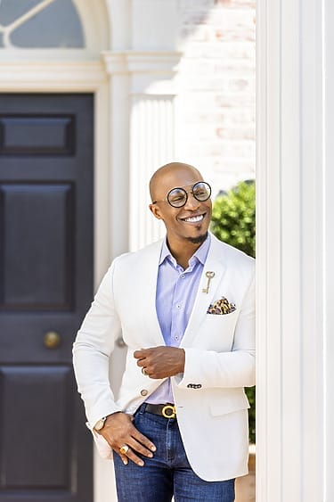 Man in white blazer and glasses smiling while leaning against a column, standing in front of a dark door with a key-shaped pin.