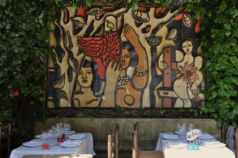 Mural with abstract figures in a leafy courtyard, surrounded by tables set for dining, featuring vibrant colors and diverse patterns.