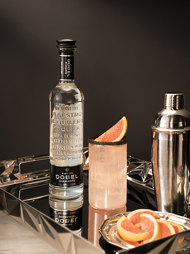 Bottle of tequila on a tray with a cocktail garnished with grapefruit slices and a cocktail shaker nearby