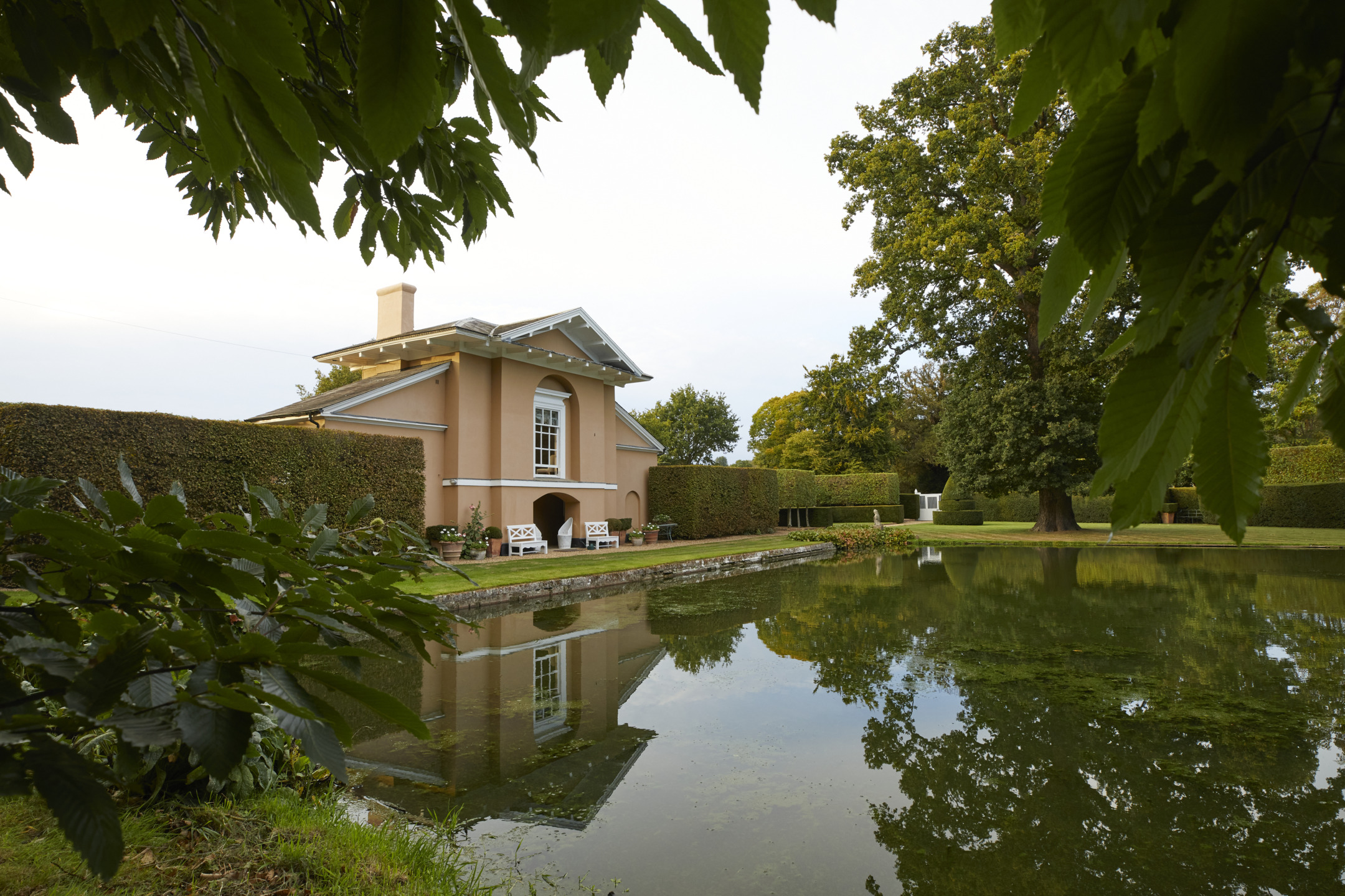 Elegant house by a serene pond, surrounded by lush greenery and framed by large leaves in the foreground.