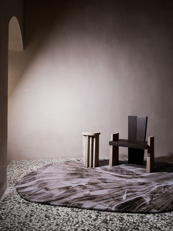 Minimalist room with a modern wooden chair, small round table, and a textured oval rug on a terrazzo floor.